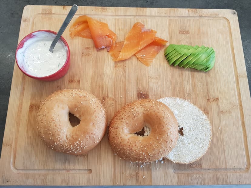 Tous les ingrédients pour la recette déposé sur une planche a découpé : suace, saumon, tranche d'avocat et pain à bagel